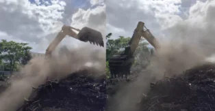Máquina escavadeira trabalhando em área com fumaça e nuvens, em meio a vegetação e entulho.