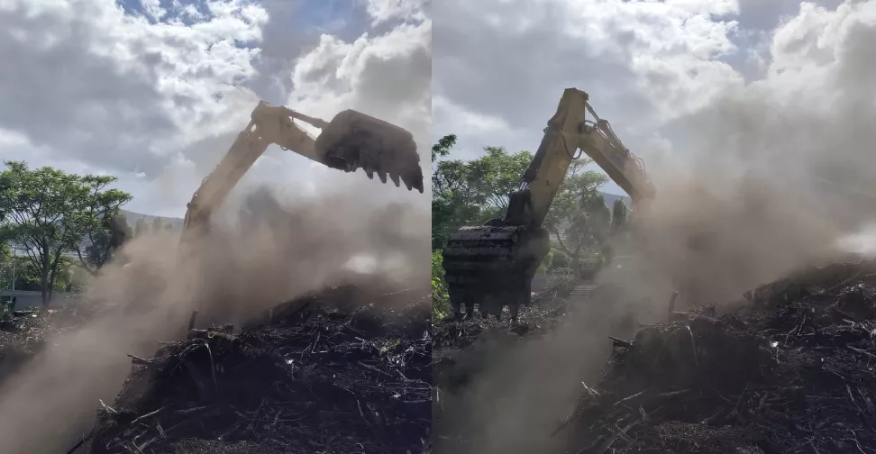 Máquina escavadeira trabalhando em área com fumaça e nuvens, em meio a vegetação e entulho.