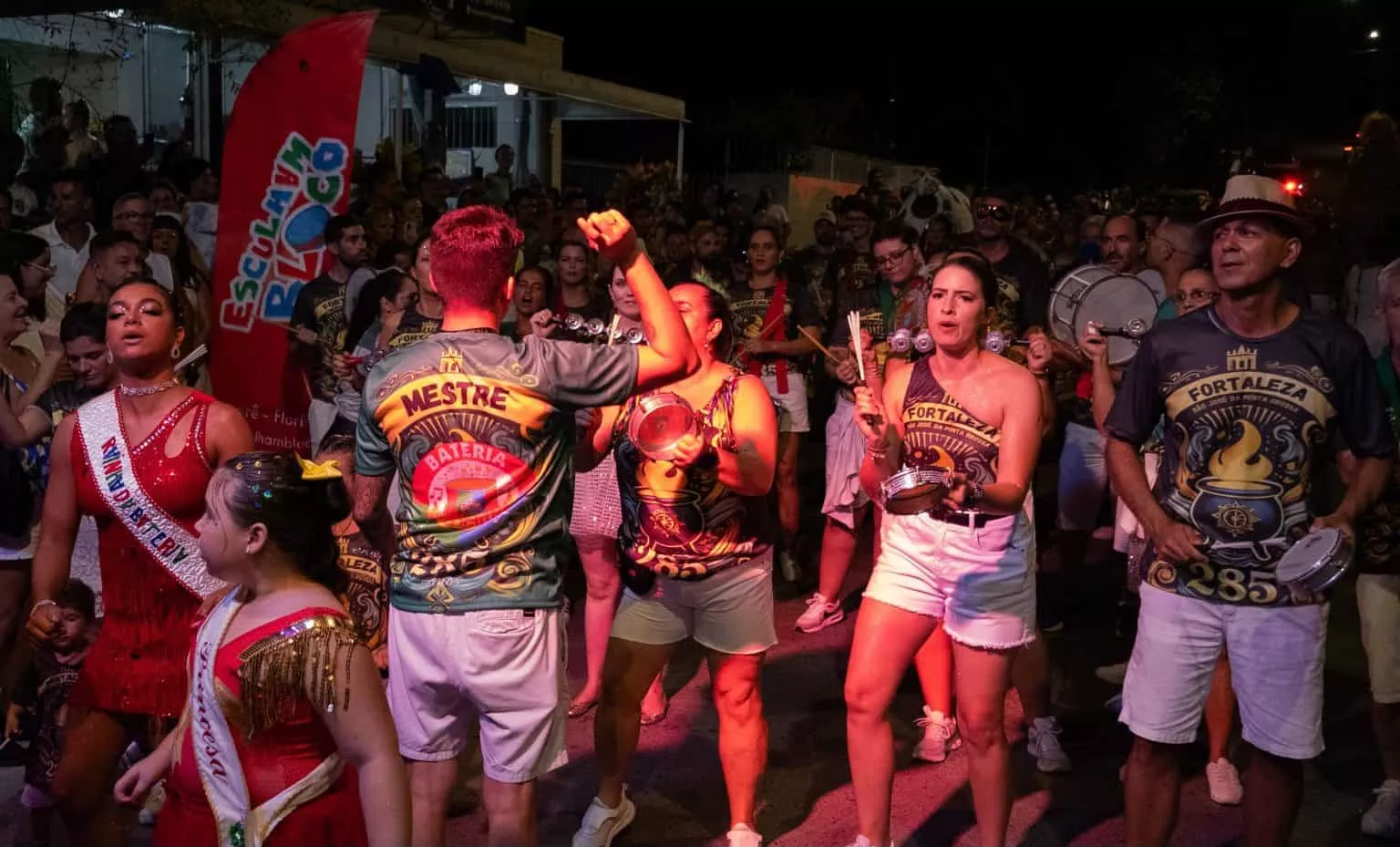 Grupo de pessoas dançando e tocando instrumentos em desfile de Carnaval, com roupas coloridas e animadas.