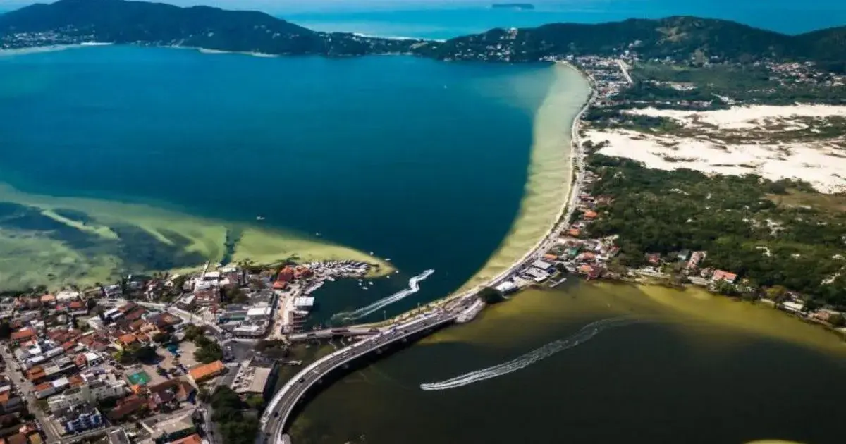 Vista aérea da Lagoa da Conceição, com águas claras e áreas urbanas ao redor, destacando a ponte e a costa.