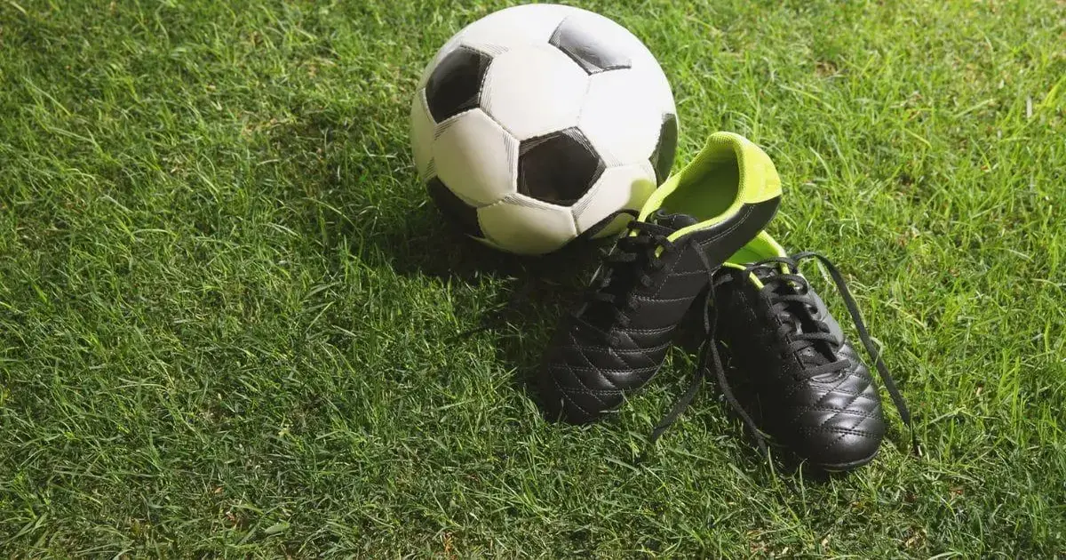 Bola de futebol preta e branca ao lado de um par de tênis de futebol pretos com detalhes verdes em um campo de grama.