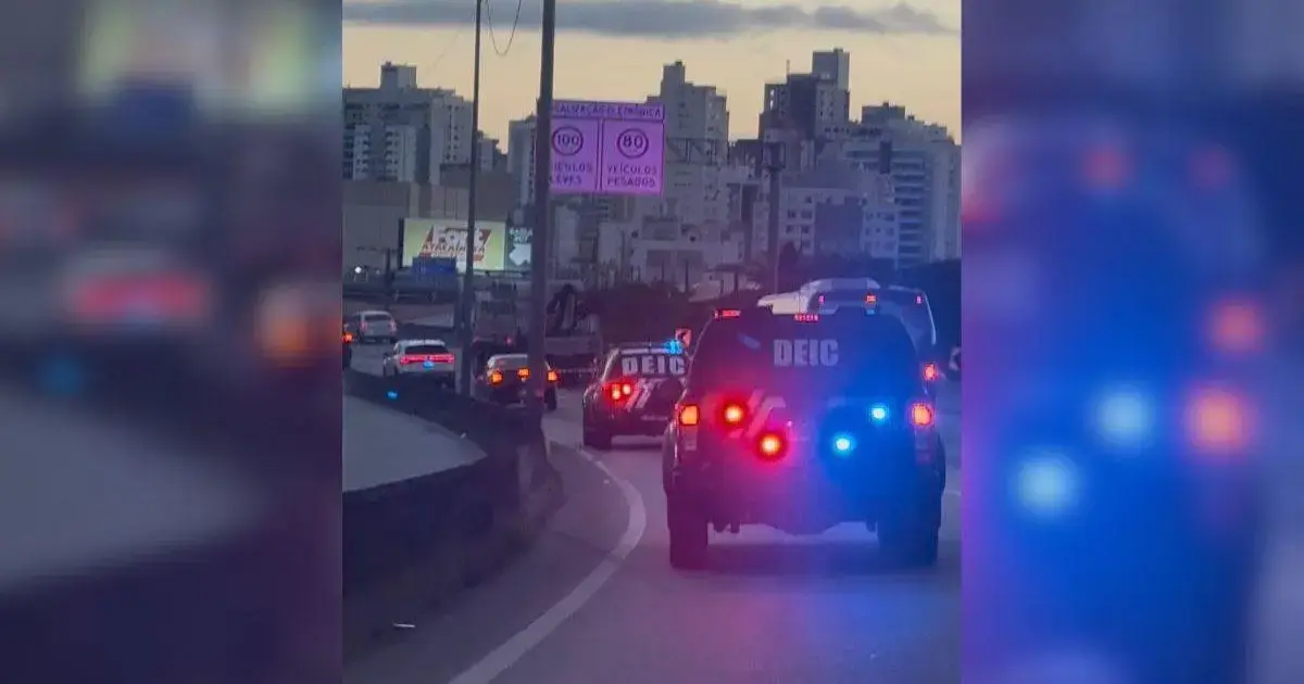 Viatura policial com luzes piscantes em estrada, prédios ao fundo e sinalização viária visível.