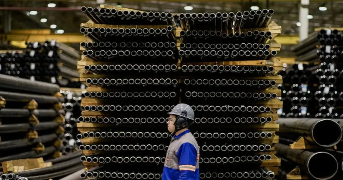 Homem com capacete em pé ao lado de pilhas de tubos de plástico em ambiente industrial.