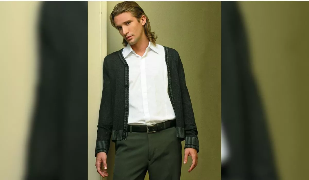 Homem com cabelo longo e liso, usando camisa branca e suéter escuro, posando em ambiente interno.