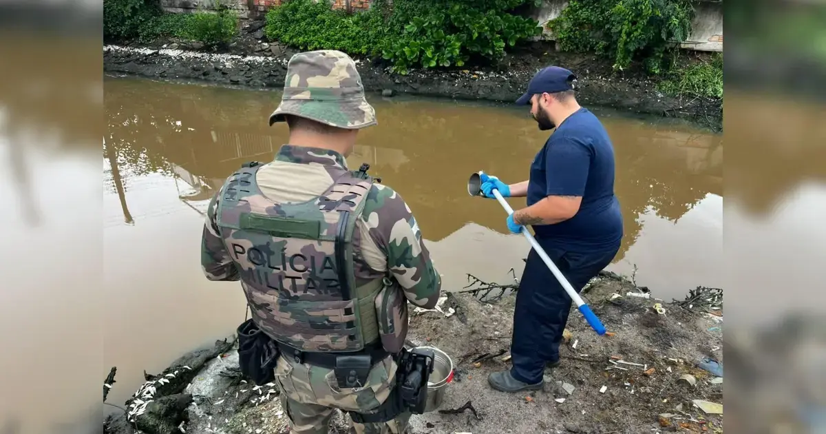 Dois homens, um de uniforme militar e outro com luvas, analisam a água turva do Rio Imaruim.