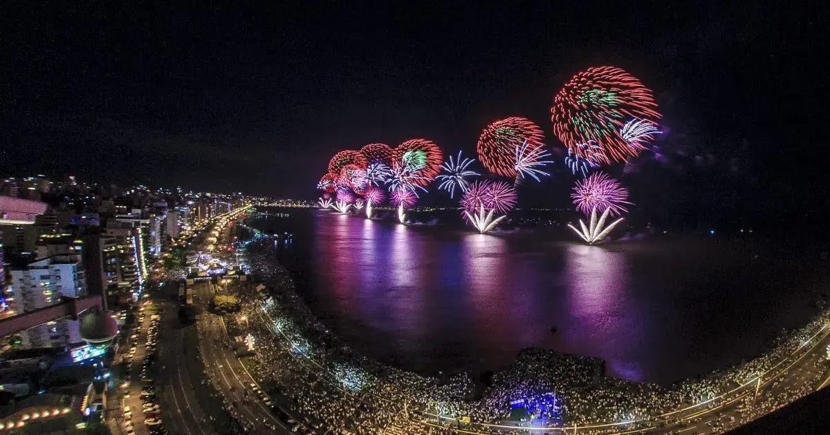 Fogos de artifício coloridos iluminando o céu noturno sobre um rio, com reflexos na água e multidão ao fundo.