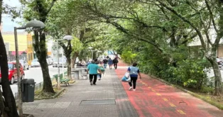 Pessoas caminhando em calçada arborizada, com ciclovia vermelha ao lado e bancos de praça.