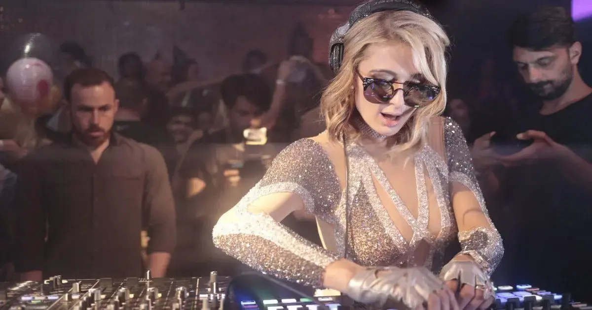Paris Hilton toca como DJ em festa, usando óculos escuros e roupas brilhantes, com público animado ao fundo.