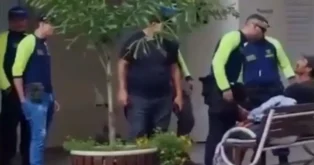Grupo de policiais em uniforme conversando com um homem em um banco, em ambiente urbano.