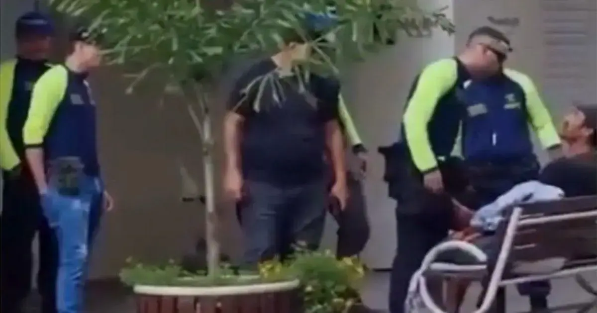 Grupo de policiais em uniforme conversando com um homem em um banco, em ambiente urbano.