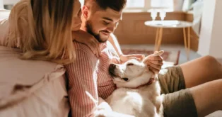 Casal acariciando um cachorro deitado entre eles, em um ambiente aconchegante e iluminado.