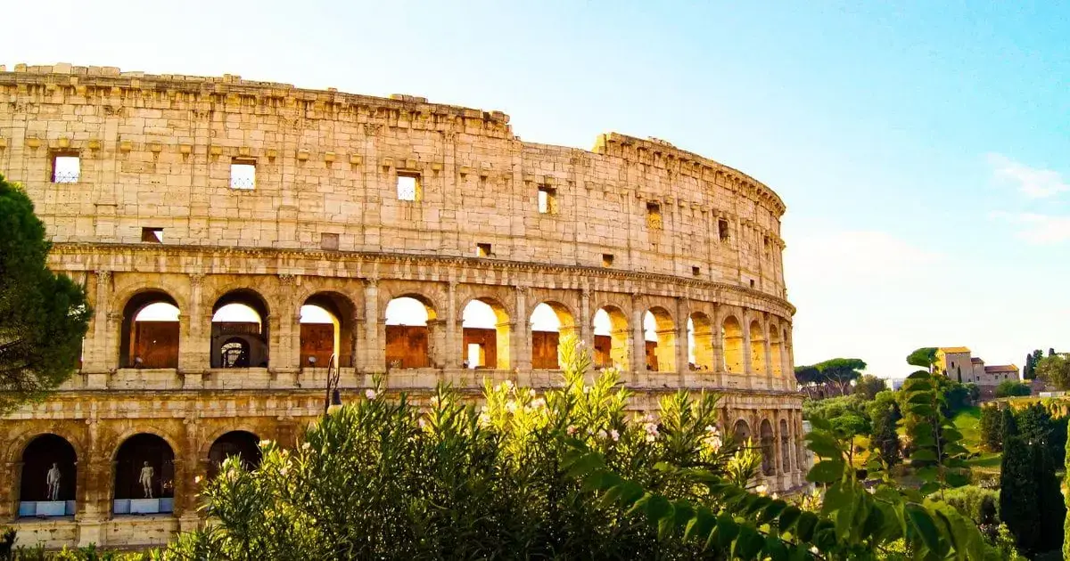 Coliseu de Roma cercado por vegetação, sob céu azul e iluminado pelo sol.