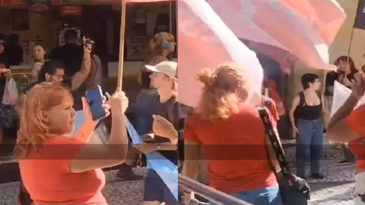Mulher com cabelo laranja segura bandeira em protesto, pessoas ao fundo com cartazes e bandeiras.