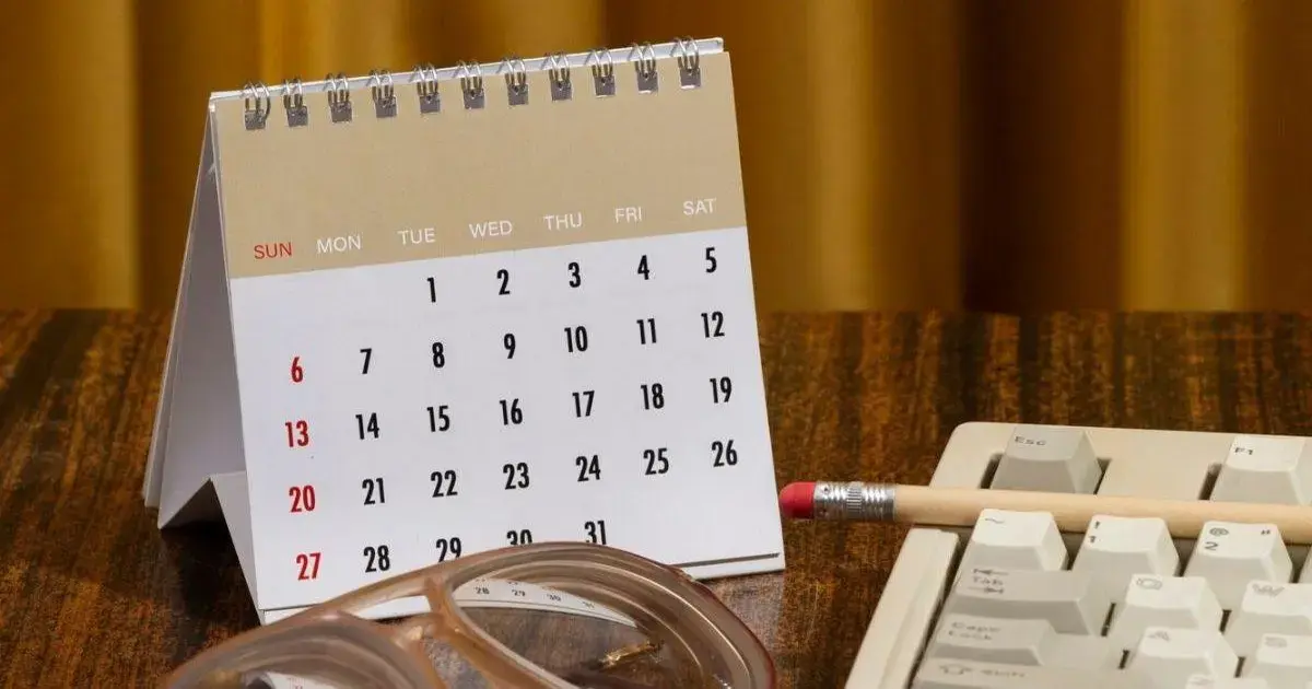 Calendário de mesa com datas destacadas, óculos e um lápis sobre uma mesa de madeira.