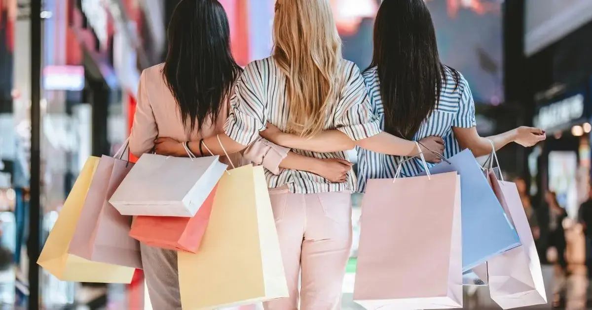 Três mulheres de costas, segurando sacolas de compras coloridas, em um shopping moderno.