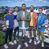 Cinco homens sorrindo em frente a um resort, usando roupas coloridas e tênis brancos, com paisagem ao fundo.