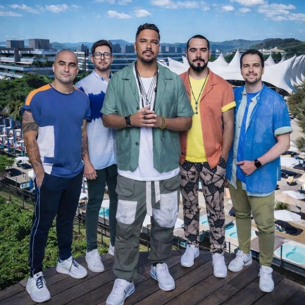 Cinco homens sorrindo em frente a um resort, usando roupas coloridas e tênis brancos, com paisagem ao fundo.