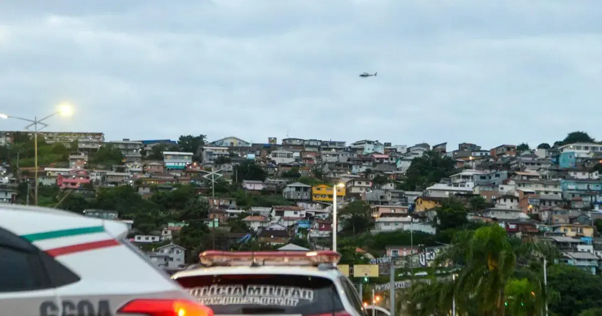 Helicóptero sobrevoa área urbana com casas coloridas em encosta, viatura da polícia em primeiro plano.