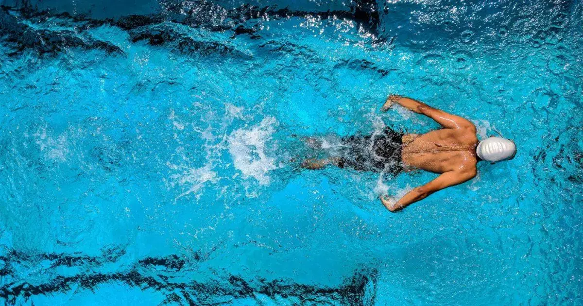 Homem nadando em piscina com água azul clara, usando touca de natação e nadando em estilo livre.