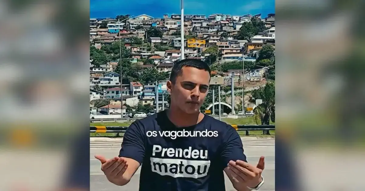Homem com camiseta preta, gesticulando, em frente a um morro com casas coloridas ao fundo.