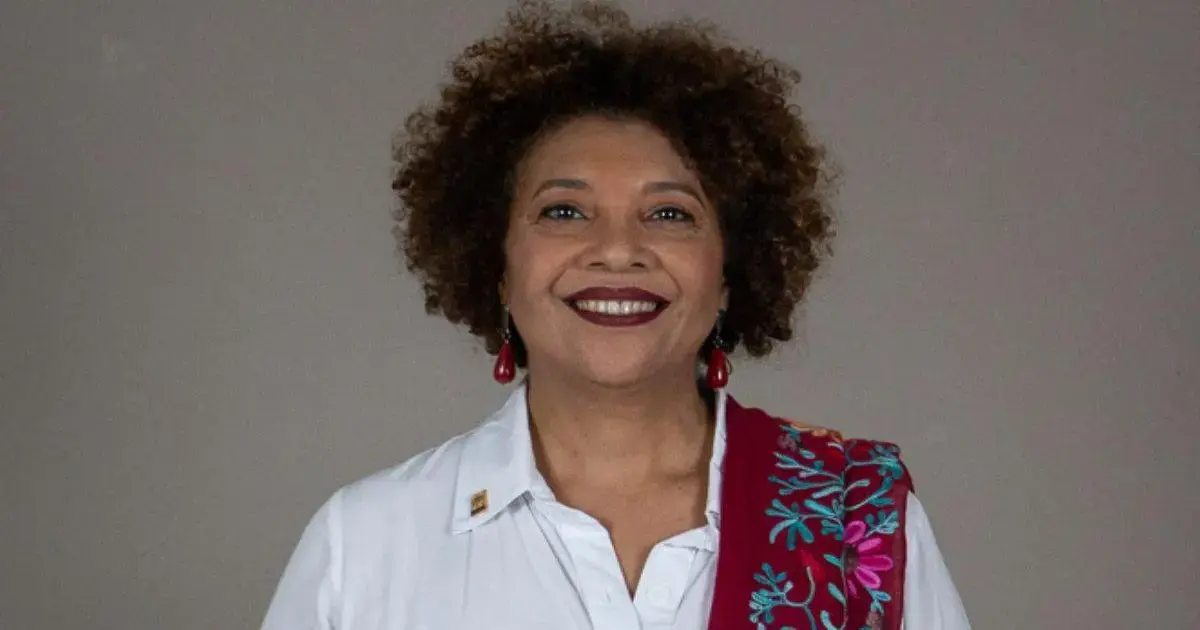 Mulher com cabelo cacheado e sorriso, usando camisa branca e echarpe colorida com bordados.