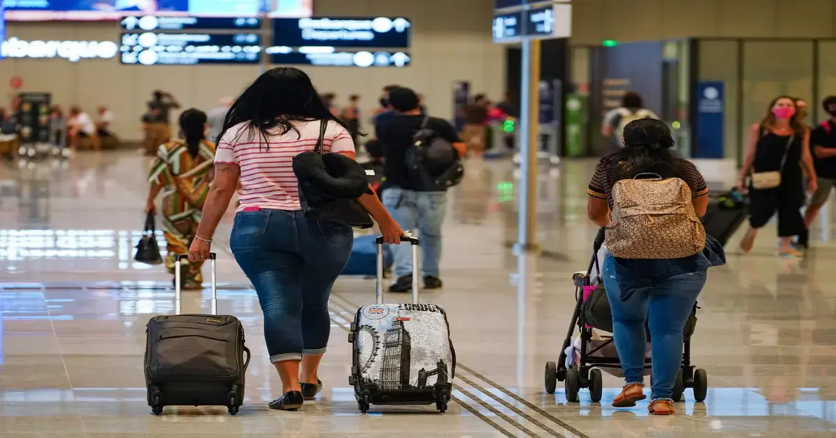 passageiras caminhando no aeroporto com malas e carrinhos de bebê, ambiente movimentado e sinalização ao fundo