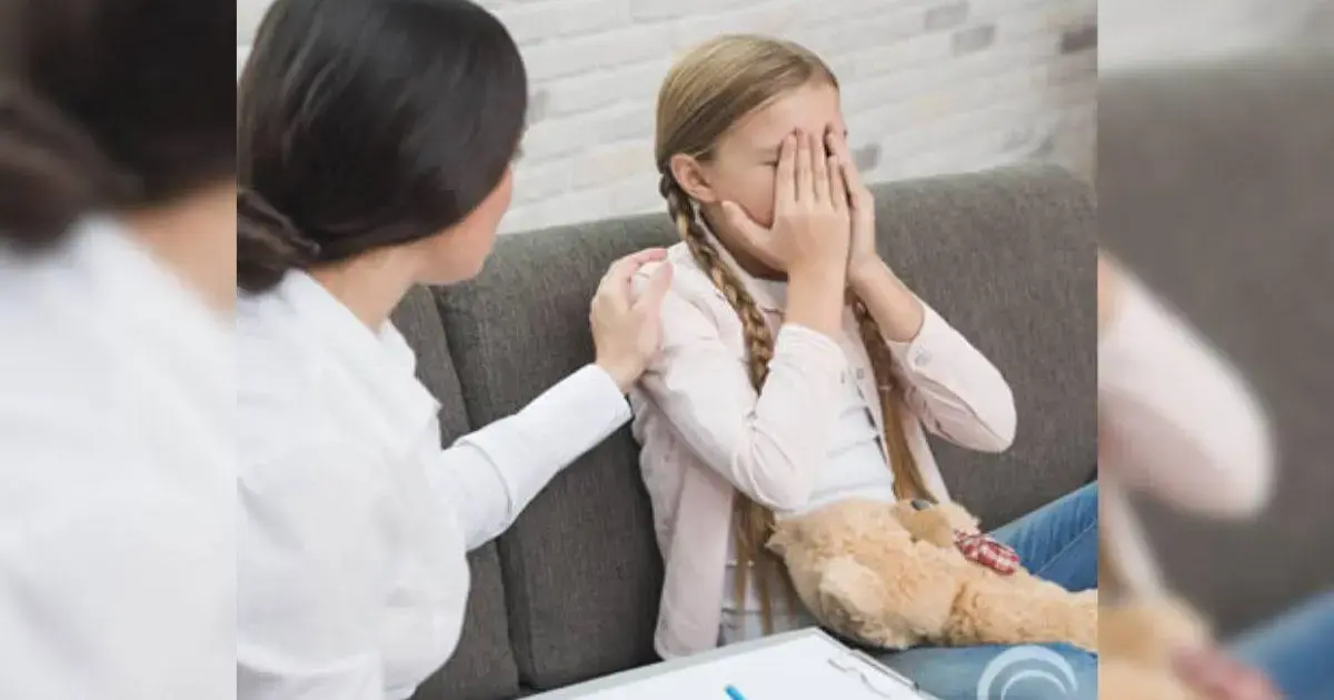 Criança com cabelo trançado cobre o rosto com as mãos, sentada no sofá ao lado de um adulto.