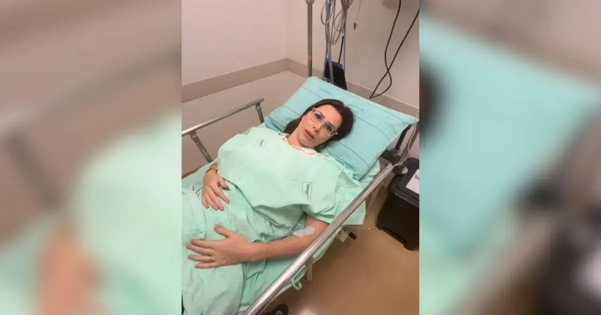 Pessoa deitada em uma cama de hospital, vestindo um roupão verde e com um semblante sério.