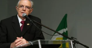 Homem de terno escuro falando ao microfone, com bandeira ao fundo e mãos sobre uma mesa de vidro.