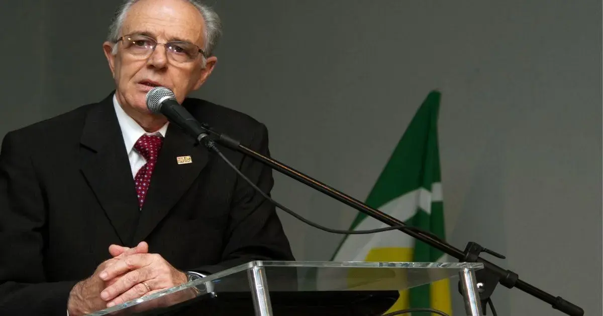 Homem de terno escuro falando ao microfone, com bandeira ao fundo e mãos sobre uma mesa de vidro.