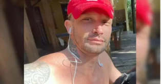 Homem com cabelo curto e boné vermelho, usando fones de ouvido, em ambiente externo de madeira.