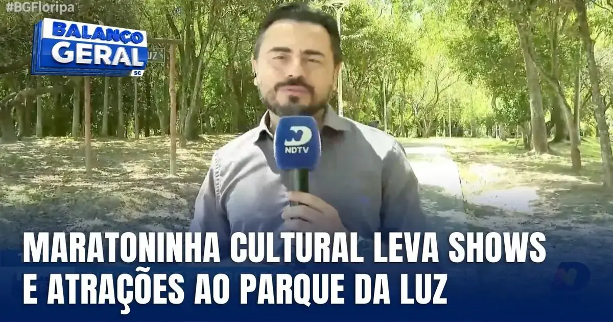 Apresentador em um parque, reportando sobre a Maratoninha Cultural, com letreiro destacando shows e atrações.