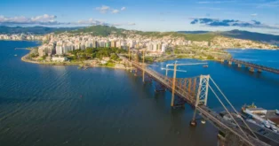 Vista aérea da cidade com prédios à beira de um rio e uma ponte de ferro em construção.