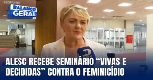 Mulher em ambiente interno, falando ao microfone, com painel informativo ao fundo sobre seminário contra feminicídio.