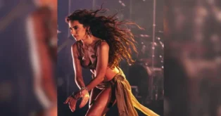 Artista em um show, movendo-se de forma expressiva, com cabelo solto e roupas de performance.