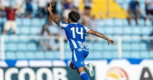 Jogador de futebol celebra um gol, com a mão levantada, vestindo uniforme azul com número 14. Torcedores ao fundo.