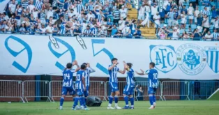 Torcedores aplaudindo e jogadores do time se cumprimentando em campo, com bandeira e placar ao fundo.