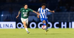 Dois jogadores de futebol em ação, um vestindo uniforme verde e branco e o outro azul e branco, em campo iluminado.
