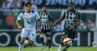 Dois jogadores de futebol em campo, um com uniforme branco e o outro com uniforme preto e listrado, ambos correndo atrás da bola.