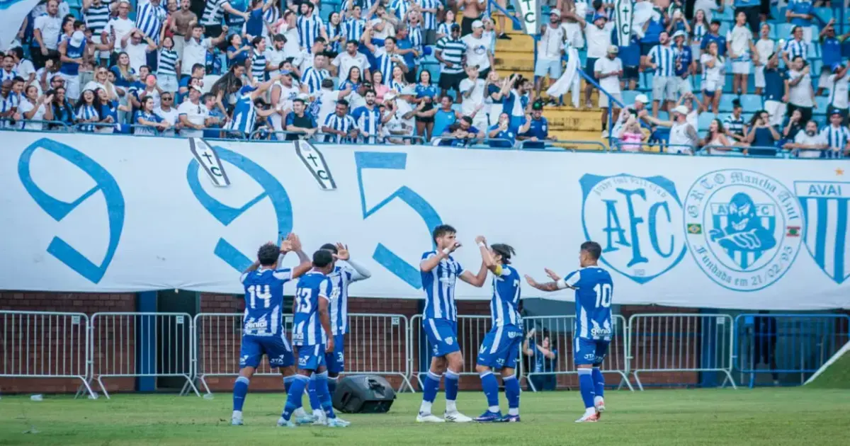Torcedores comemoram na arquibancada enquanto jogadores do time celebram em campo, com bandeiras e camisas.
