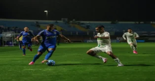 Jogadores em campo durante uma partida de futebol à noite, com um atleta em azul driblando um adversário em branco.