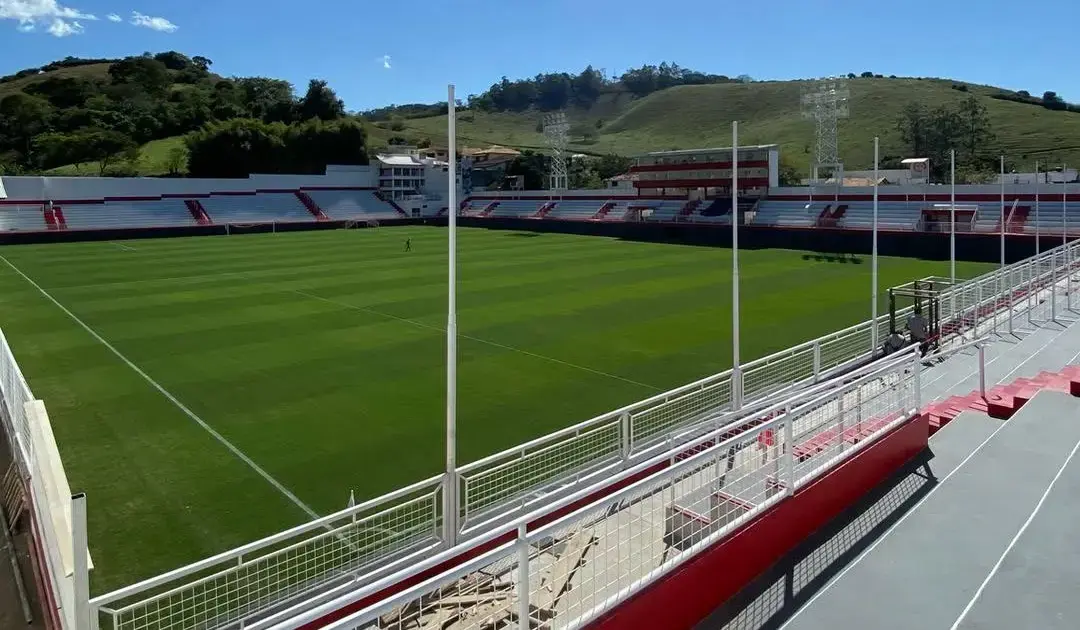 Campo de futebol em um estádio com arquibancadas vermelhas e brancas, rodeado por colinas verdes e céu azul.