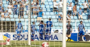 Jogadores comemoram após um gol em um jogo de futebol, com torcedores ao fundo.