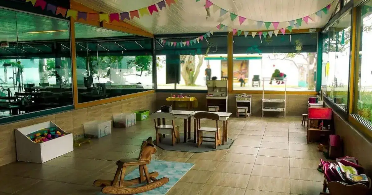 Espaço interno com mesas e cadeiras, brinquedos coloridos e decoração festiva em uma sala infantil.