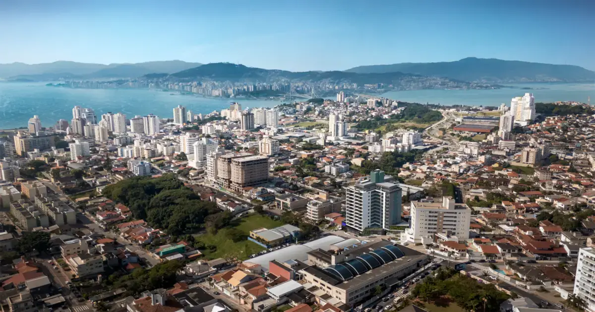 Vista panorâmica de uma cidade costeira, com edifícios, ruas e áreas verdes, ao fundo montanhas e o mar.