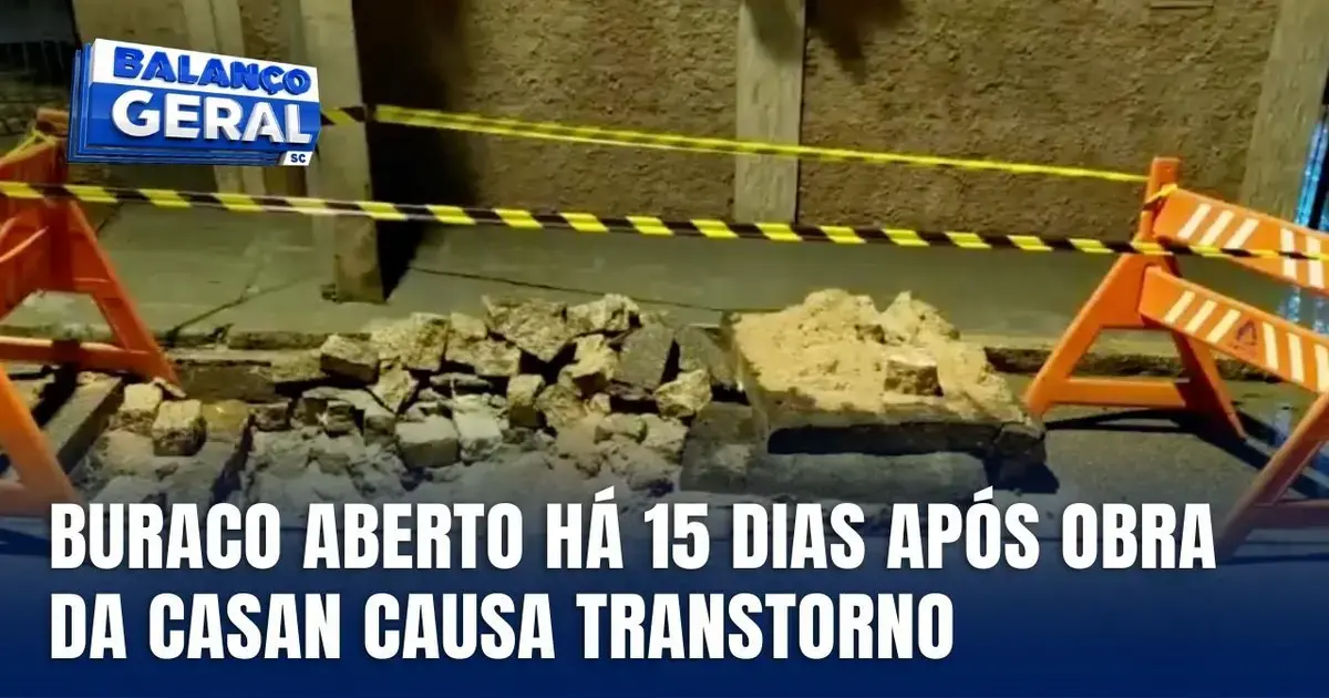 Buraco na calçada com pedras e terra, cercado por cones de sinalização e fita de segurança. Texto: 
