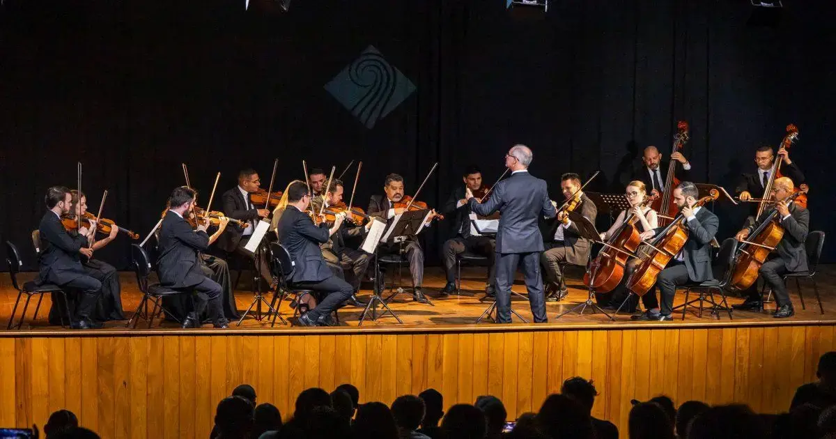 Orquestra em apresentação com músicos tocando instrumentos de corda sob a direção de um maestro em um palco.