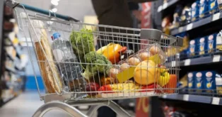 Carrinho de supermercado com frutas, legumes, ovos, garrafa de água e pão. Fundo com prateleiras de produtos.