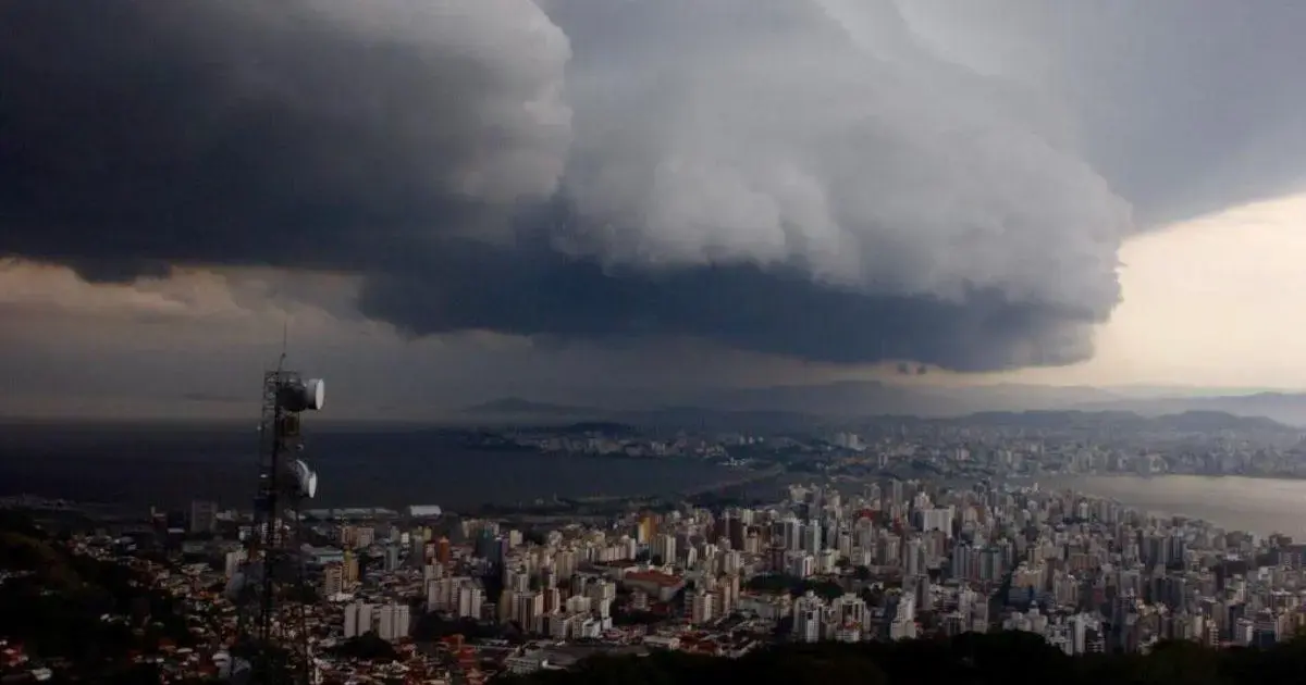 Vista de uma cidade com prédios e uma torre de comunicação, sob nuvens escuras e tempestade iminente.