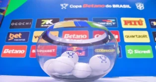 Recipiente de vidro com bolinhas brancas dentro, ao fundo, banners de patrocinadores e o logo da Copa Betano do Brasil.